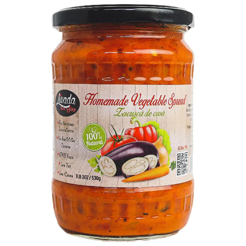 Clear glass jar filled with bright orange-red vegetable spread and sealed with a black screw-top lid. The front label displays the Lunada logo and the product name 'Homemade Vegetable Spread.' Illustrations of eggplant, tomatoes, red pepper, onion, and carrot appear on the label, along with a '100% Natural' badge and claims such as GMO free, no artificial colors, and low fat. The jar contains 530 g (1 lb 2 oz) of spread